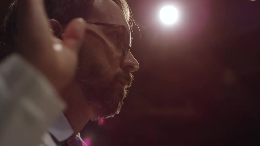 Bearded man wearing sunglasses holds his hand up while talking