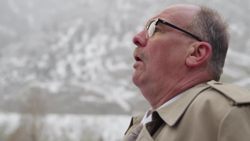 Elderly family doctor wearing glasses stands outdoors while snowing