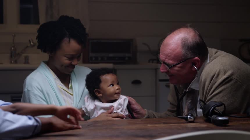 A mother holds her baby in her lap as a doctor examines her during a home visit