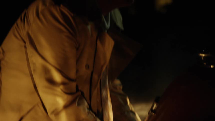 A man in a trench coat opens the rear hatch of a vehicle outdoors at night