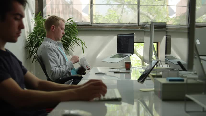 A young man sits at his desk and eats and looks at his computer while his coworker types on his keyboard