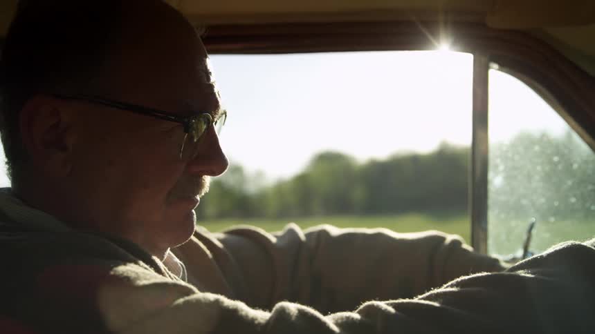 Elderly man with a trimmed mustache and glasses drives on a road in the countryside