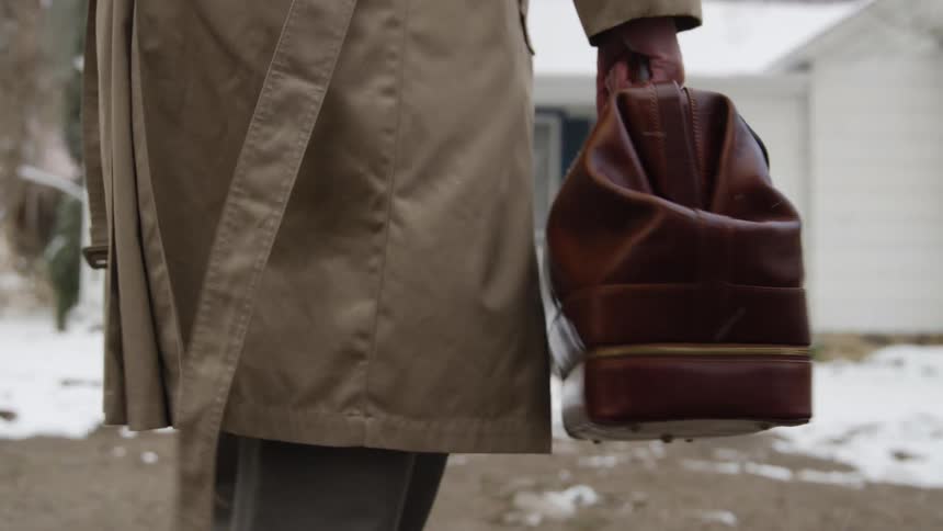 A family doctor with a trench coat and brown bag walks to a house