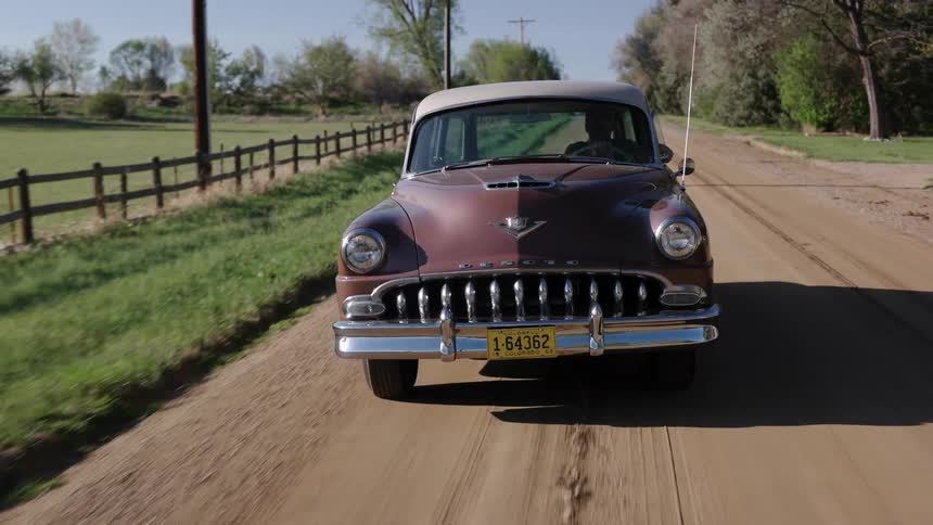 Man drives a dark red classic car on a dirt road in the countryside