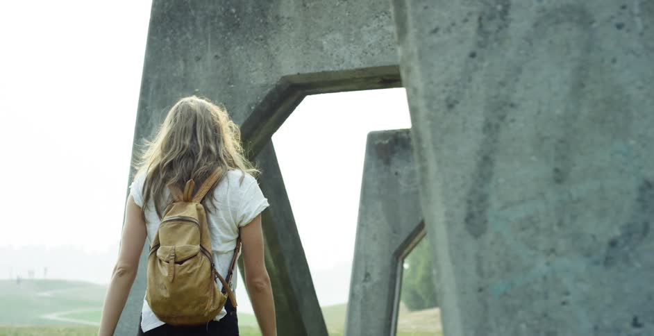A woman carrying a book walks under arched cement supports on a hillside trail