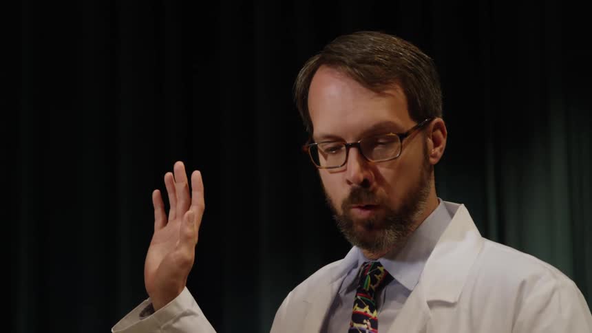 A man in a white lab coat stands on a stage and raises his left hand to take an oath
