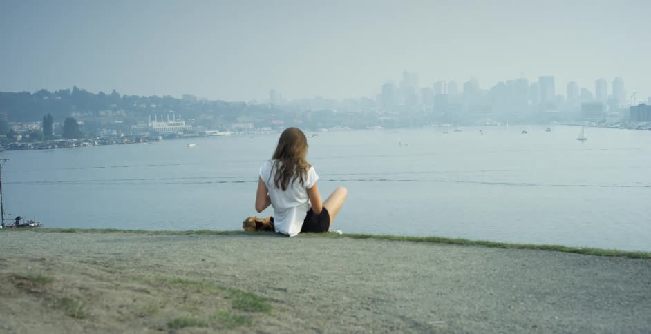 Woman with black shorts and a white shirt reads a book on a hill with tall buildings across the sea in the background
