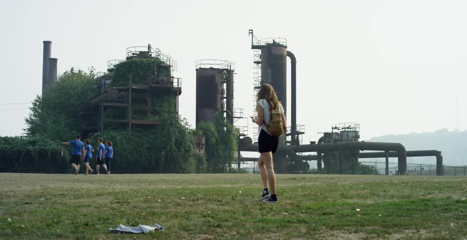 A group of kids walk past a refinery and a female teenager walks behind them on her own