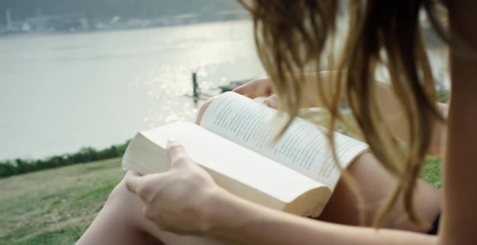 Woman with brown curly hair and black shorts sits on a hill slope and reads a book