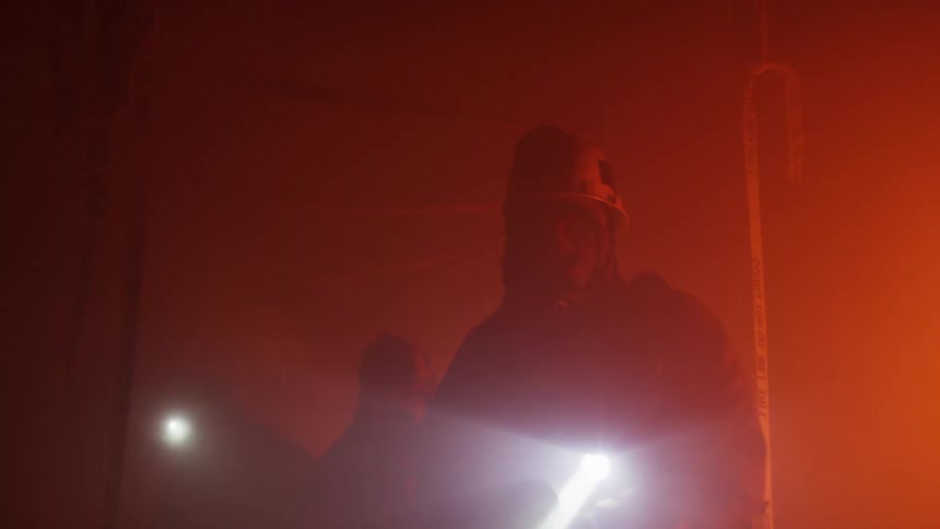 A group of firefighters in firefighting equipment moving through a dark smoky building with the help of flashlights