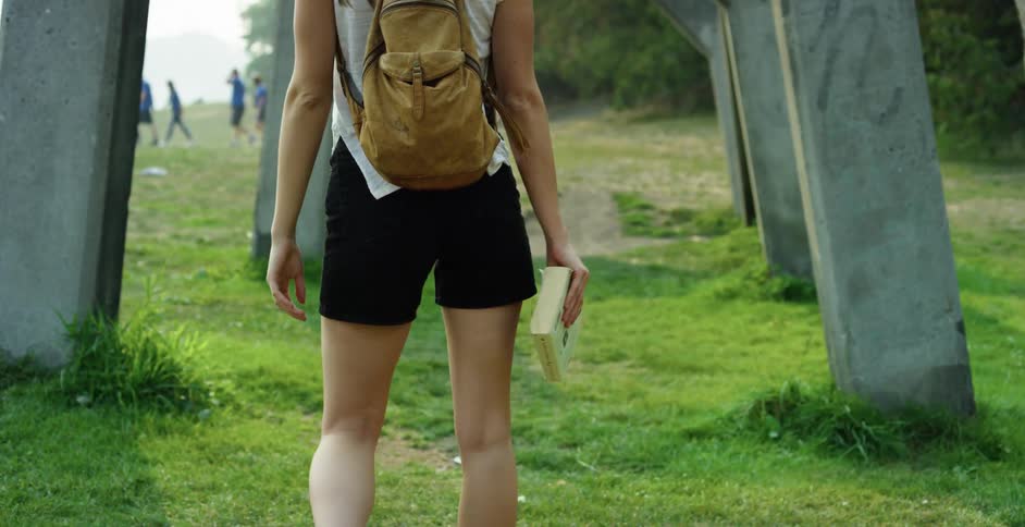 A young girl walks under a bridge carrying a book as a group of boys walk off in the distance