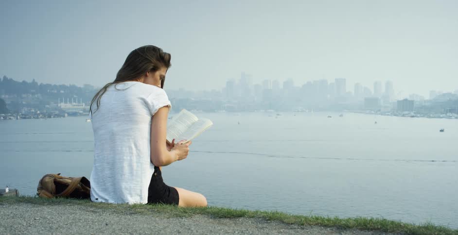 Woman with black shorts and a white shirt reads a book on a hill with high rise buildings across the sea in the background