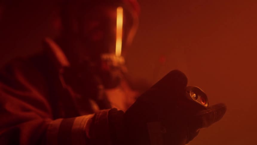 Firefighter in protective gear sweeps area with sensor as fire reflecting off face shield tints everything orange
