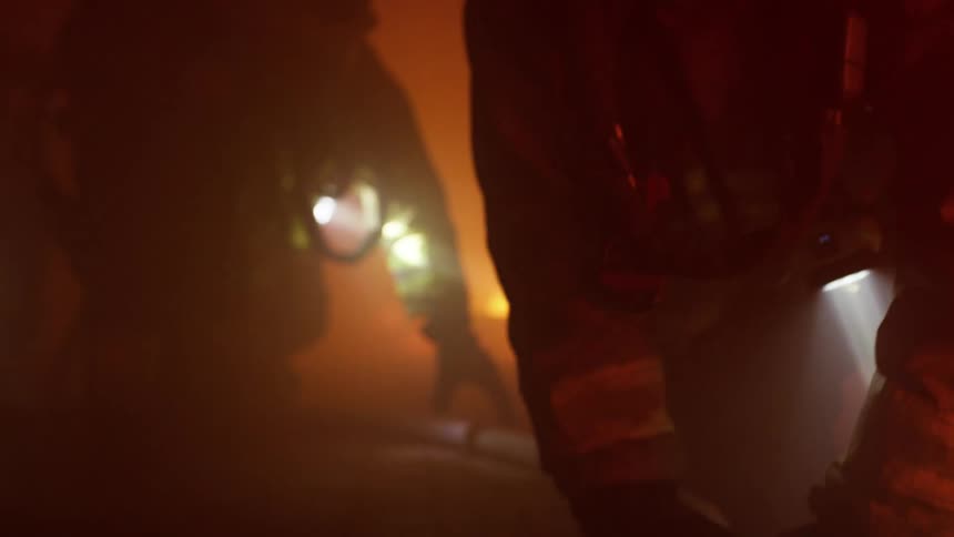 Several firefighters crawl through a smoky building using flashlights to light the way