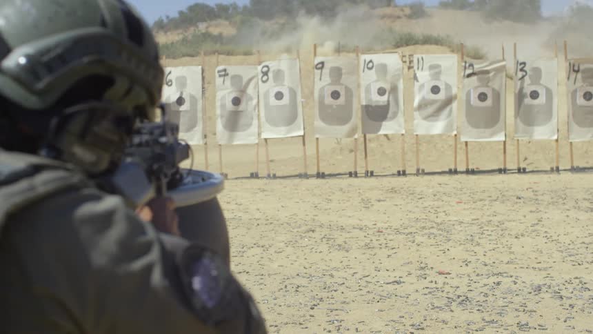 A police officer practices shooting a long gun at a line of paper targets at an outdoor range