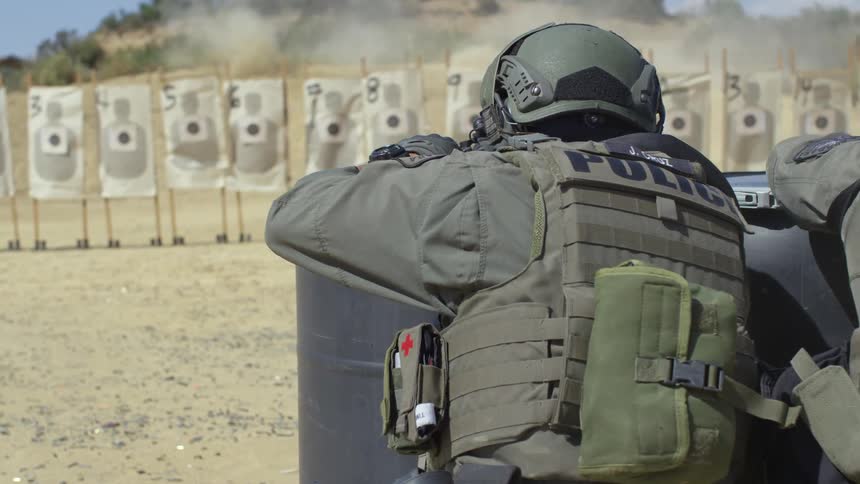 Police officers hide behind barrels and practice shooting at a line of paper targets