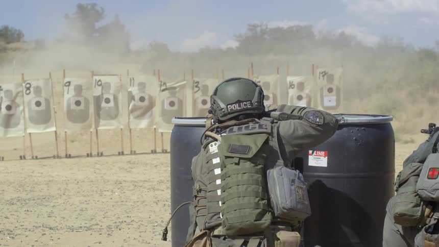 Police officers in tactical gear fire rifles at numbered targets causing casings and dirt to fly up and around