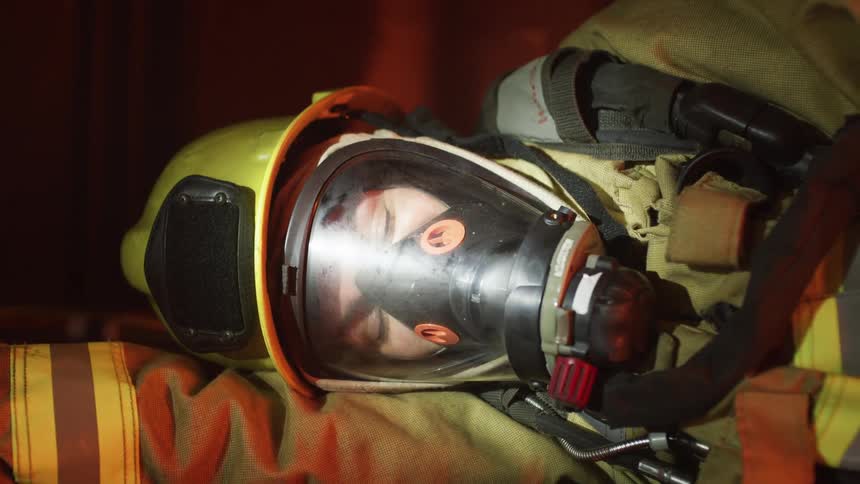 A firefighter in full gear lies on the floor unconscious as a flashlight shines on his face