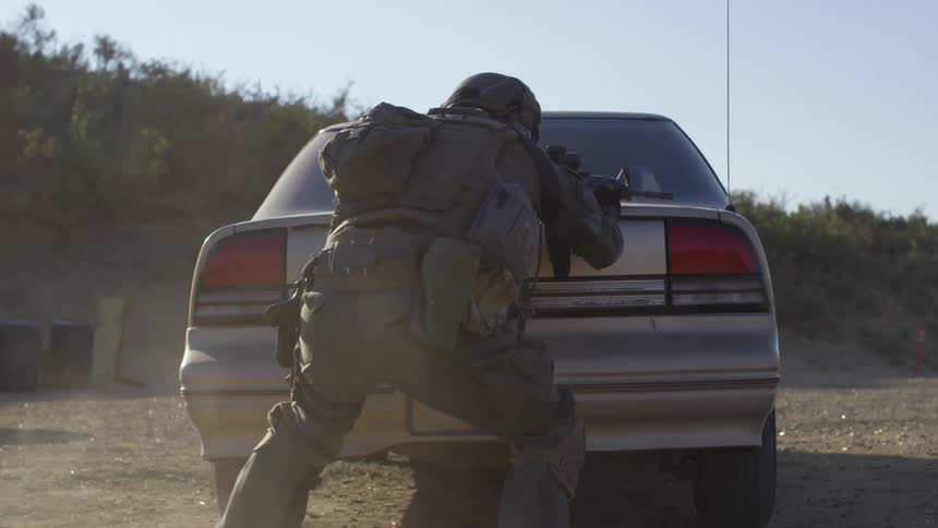 A military police runs and ducks behind a car as they shoot their gun