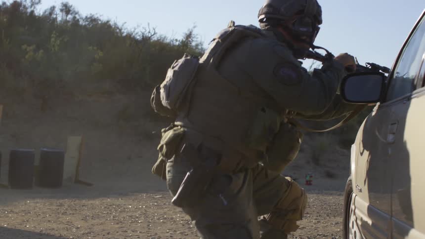 Police officer in military gear crouchs behind a car to take cover during a training exercise