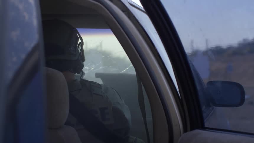 A soldier sitting in a car shoots his gun out the windshield