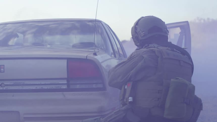 A soldier shoots his gun as he hides behind a car