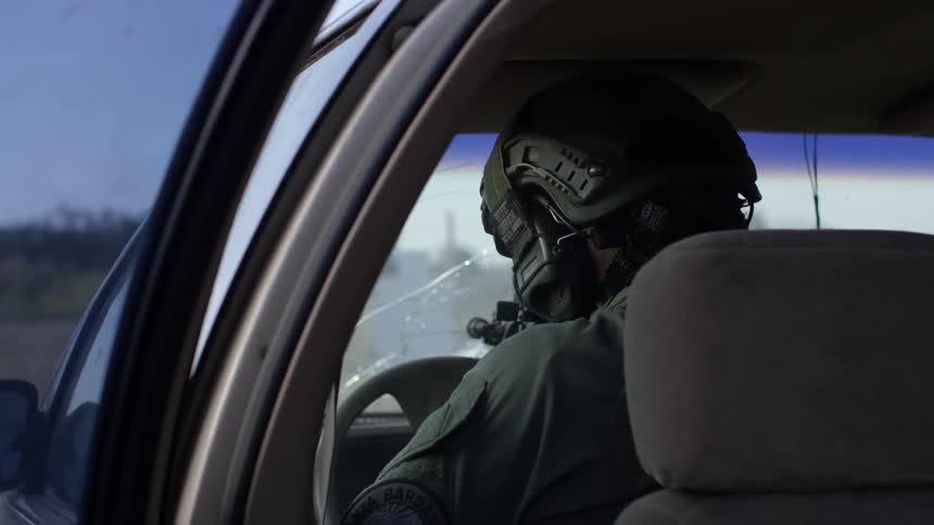 A soldier sitting in a car shoots his gun out the windshield