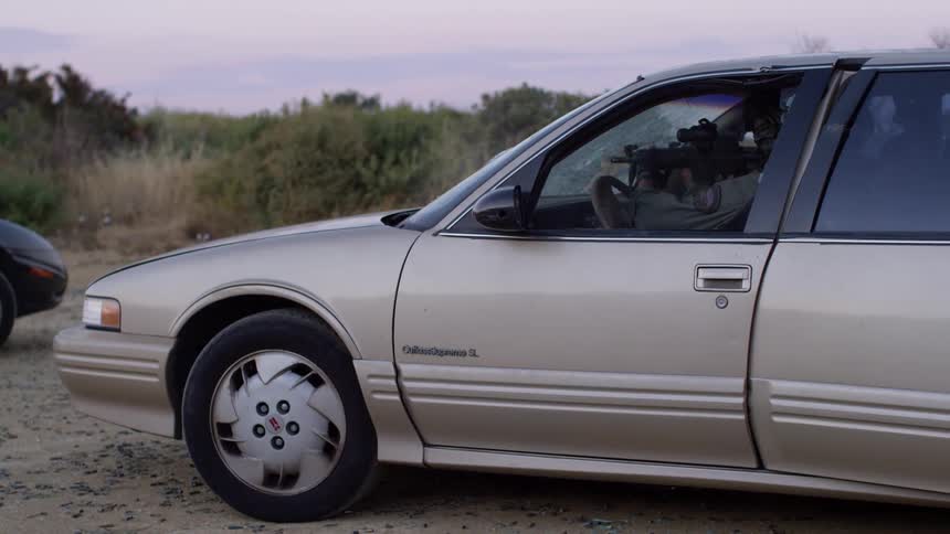 A soldier sits in a car shooting through the windshield with a rifle as another soldier exits the backseat