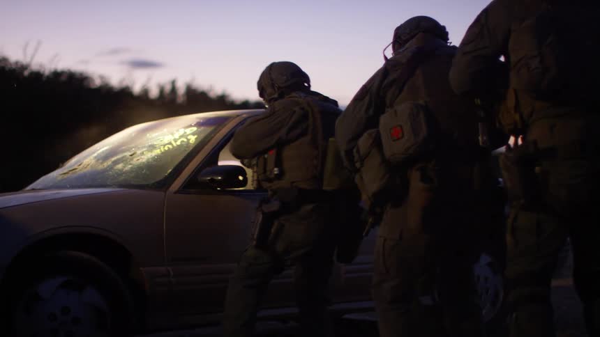Camouflaged police officers in combat armor carefully approach a damaged vehicle and fire upon the paper target within