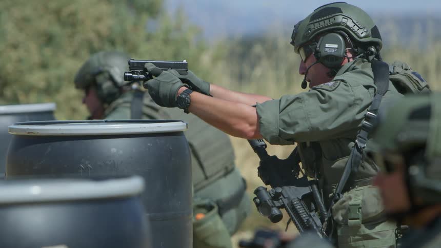Three police officers in tactical gear take aim with guns over barrels