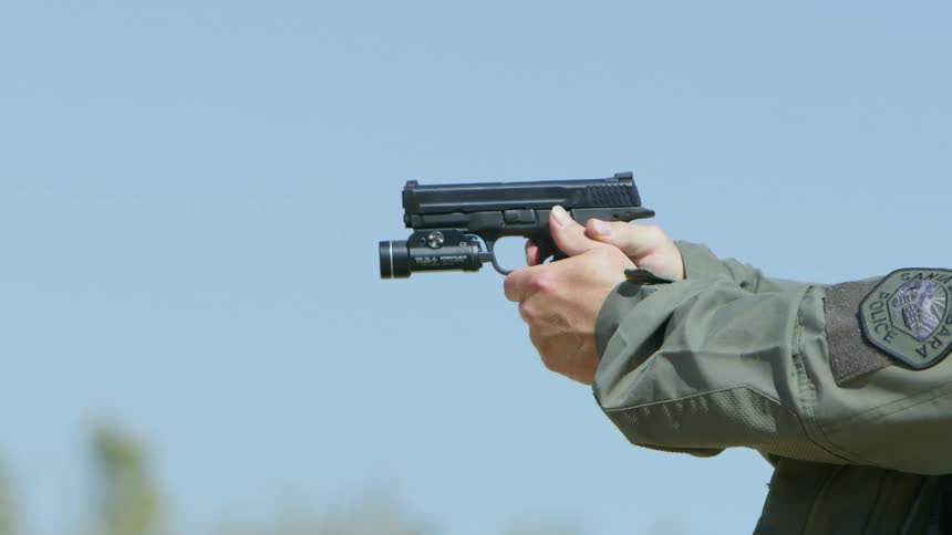 A bullet flies up and backward as a police officer fires a handgun outdoors