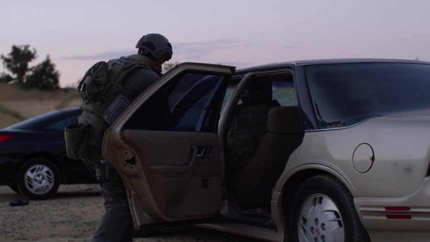 Casings fly and dust billows as a police officer in tactical gear shoots at targets while another gets out of a car