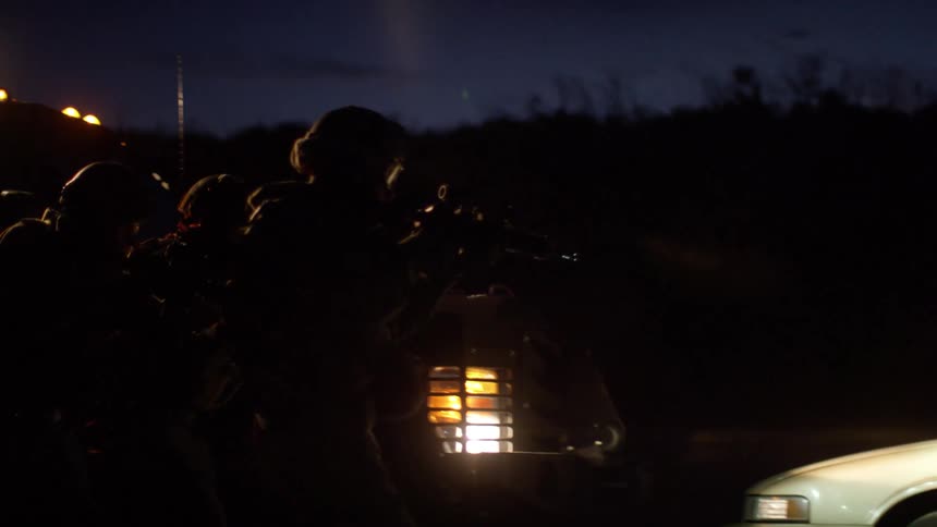 Men dressed in black body armor and helmets retreat from cars aiming rifles