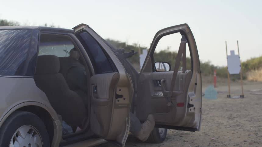 Military personnel sits in a car while firing a rifle at a target in a shooting range