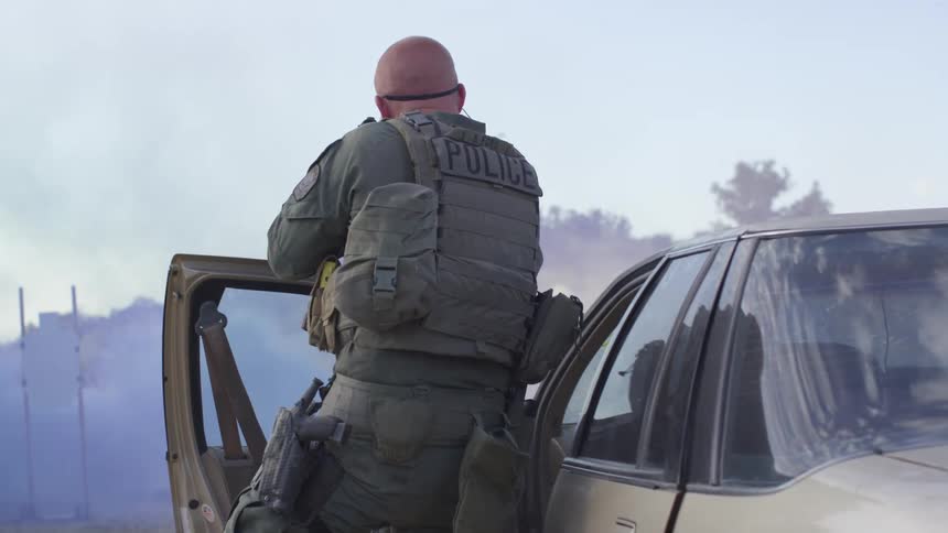 A police man in tactical gear carrying a rifle gets out of a vehicle and immediately starts shooting towards a smokey area