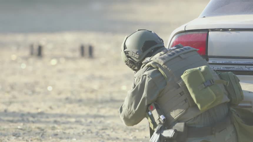 A soldier in uniform fires rounds from a weapon while hiding behind a car