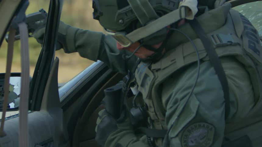 A police officer in tactical gear fires a hand gun from a car and then exits the vehicle