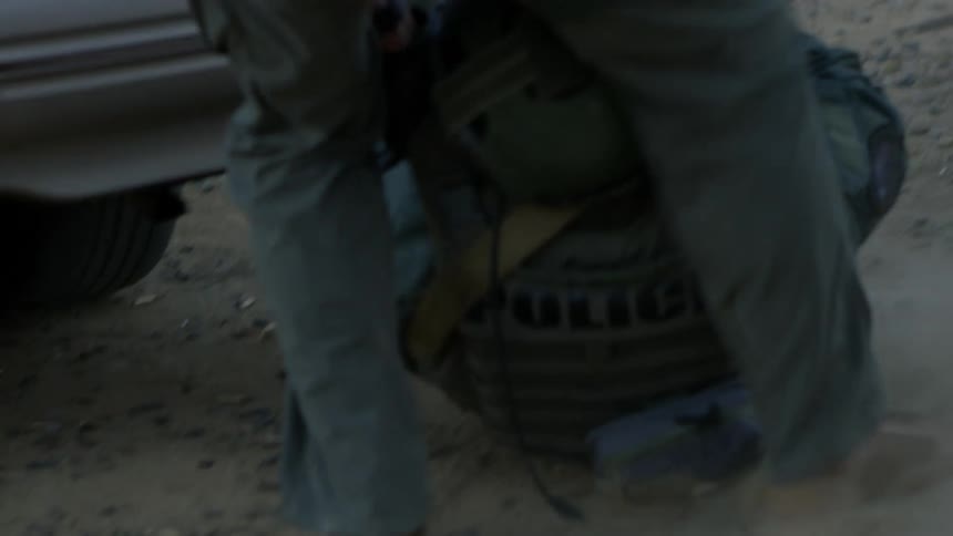 A military person drags another military person on the ground by a vehicle in a rural setting