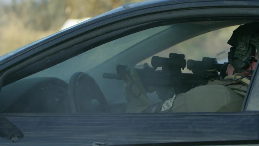 Two military police sit in front of a vehicle and shoot their guns out the front window