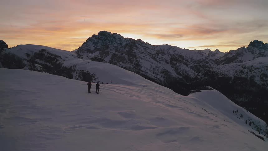Two photographers standing in snow takes photographs of snow capped mountains against the red sky