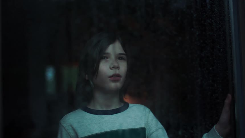 A young boy stands at a window and watches the rain outside in awe