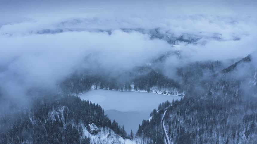 A frozen lake in a forest below a mountain shrouded in cloud and fog