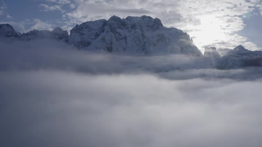 Snowy craggy mountain stands above some clouds while sun is obscured by others