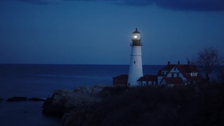A spinning light shining atop a lighthouse above a rocky cliff at night