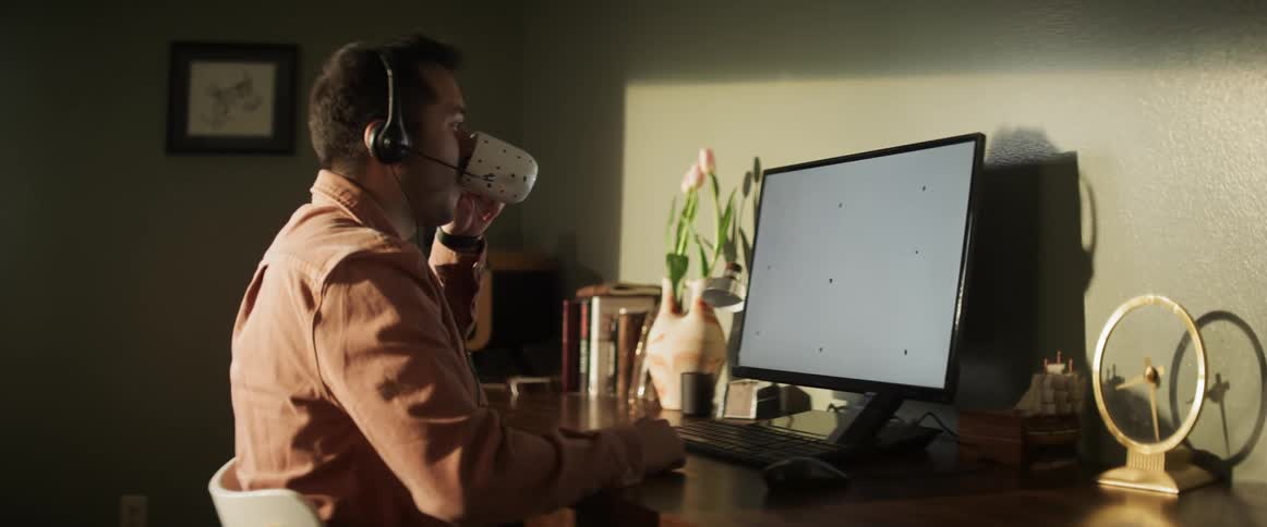 A white male sits at a computer with a headset drinking out of a mug