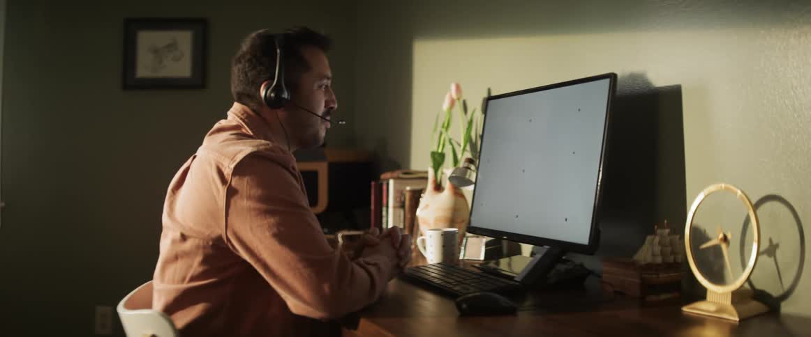 A man sits at a computer desk working and wearing a microphone head set
