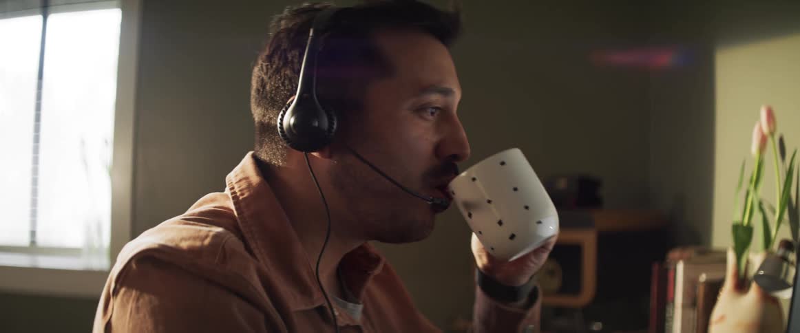 A man with a headset in front of a computer drinking tea