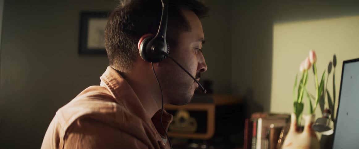A man with a headset on sits in front of his computer talking and drinking out of a mug