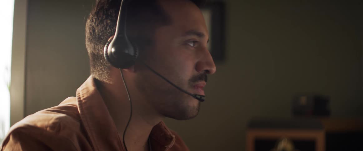 A man with a headset on talks and drinks out of a mug