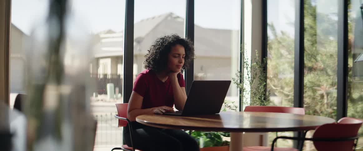 A woman sits at a table in a window lined office while working on her laptop